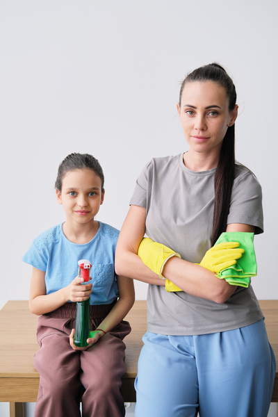A girl with her hair gathered holding detergent in a spray bottle sits on the table next to mother in yellow rubber gloves with green microfiber in her hand