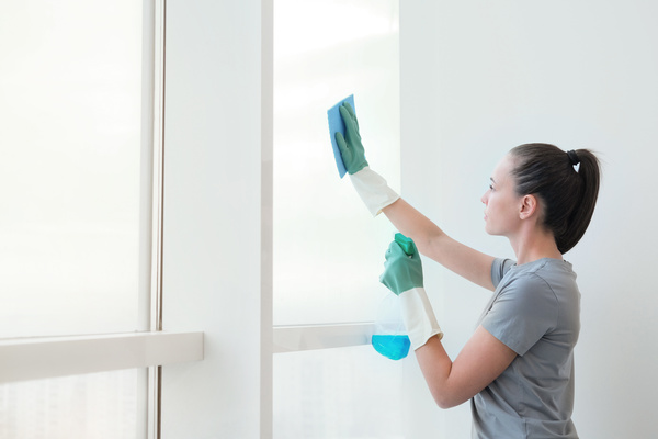 A woman in green and white rubber gloves washing a window with a blue cleaning cloth and cleaning chemicals from a spray bottle