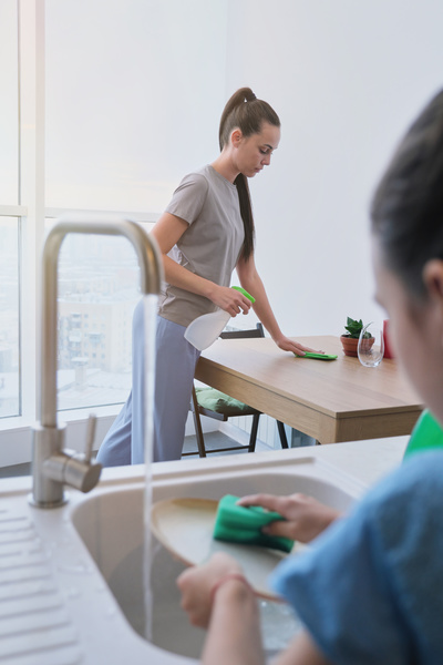 A woman with her hair in a ponytail washes the surface of the dining table with a green cleaning cloth with a spray cleaner
