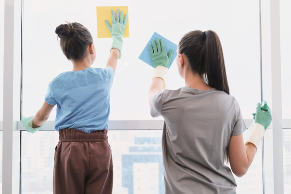 A woman with her daughter with their hair gathered in rubber gloves wash the window with sponge wipes for cleaning blue and yellow colors
