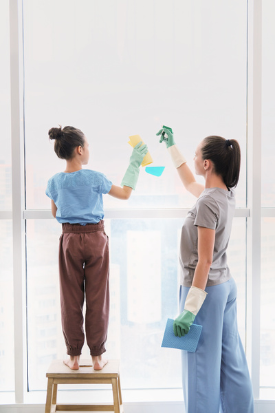 A woman applies detergent next to her daughter in green rubber gloves wiping the window with sponge wipes for cleaning blue and yellow colors