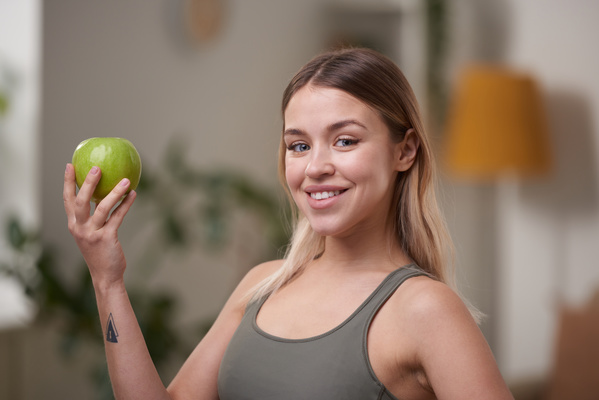 A sporty woman with dyed hair dressed in a sports top smiling to the teeth holds a green apple in her hand