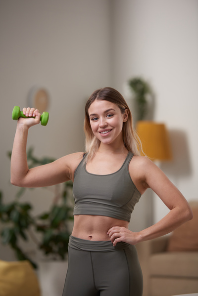 A sporty woman with dyed hair dressed in a tracksuit smiling to the teeth holds a green dumbbell in her hand