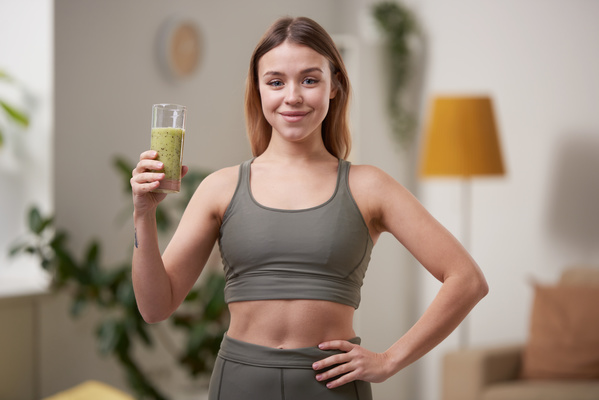 A woman of athletic build with dyed hair dressed in a tracksuit stands with a glass of green smoothie