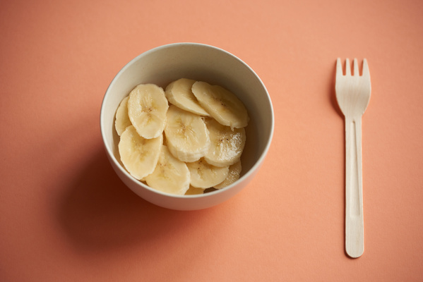 A white bowl with banana slices with a plastic fork on a peach-colored surface