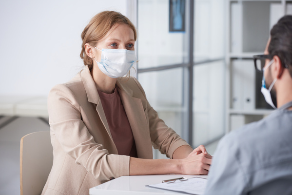 A patient in a medical mask sitting opposite the doctor tells him about her complaints