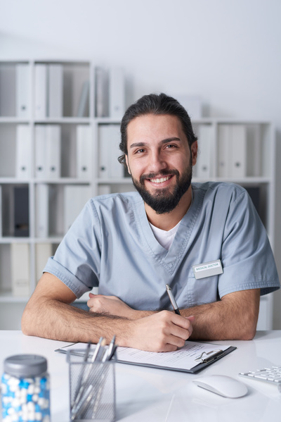 A male doctor with a beard sitting in a bright office at a desk with a document on a clipboard holding a pen and smiling