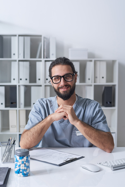 A doctor with a beard and glasses sitting in a bright office at a desk with his hands on his elbows and smiling