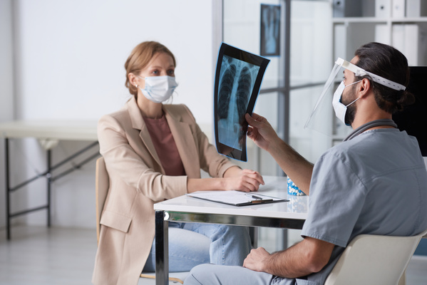 A male doctor in medical protection on his face looks at the results of an X-ray of the patients thoracic region while sitting with her at a desk in the office