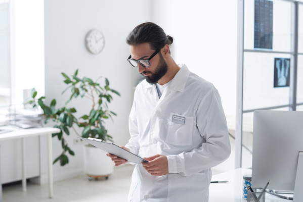 A male doctor in a white robe and glasses intently studies documents on a clipboard standing in the office