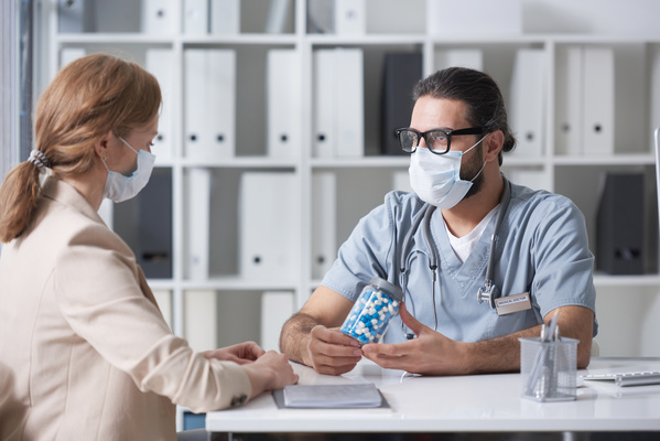 A doctor in uniform sitting at a desk prescribes a medicament in blue and white capsules to a patient in a medical mask