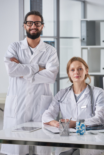 A medic in glasses and a white robe stands with his arms crossed next to the table where his colleague sits with a phonendoscope around her neck