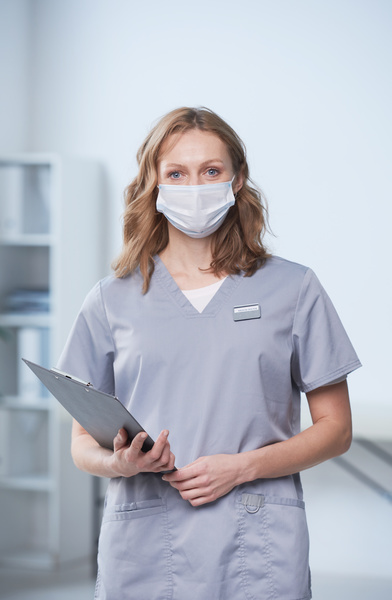 A female physician with brown curly hair and a medical mask on her facedressed in a gray medcal uniform holding a clipboard