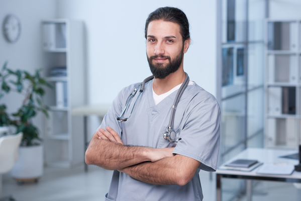 A male doctor with a thick beard in uniform and with a phonendoscope crossed his arms over his chest standing in the office