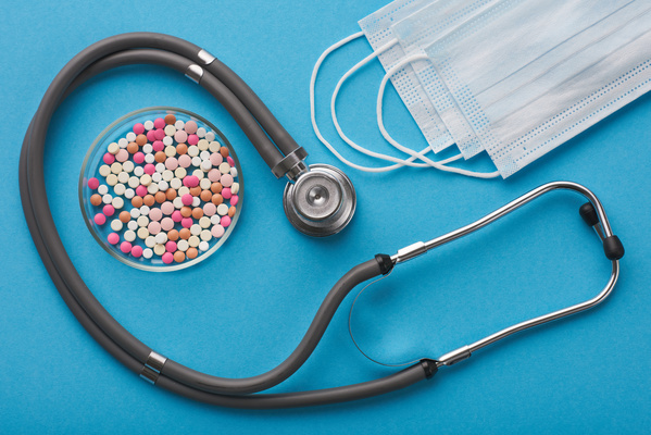 Stethoscope with multicolored pills in a small glass container and with medical masks on a blue surface