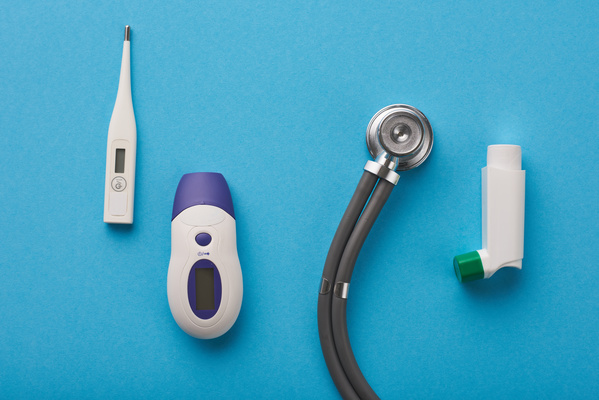 Medical equipment such as an electronic thermometer a blood glucose meter a stethoscope head and an inhaler is on a blue surface.