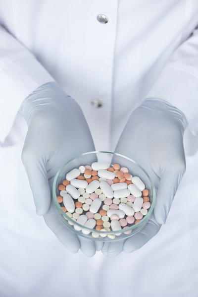 A round glass petri dish with pills of different shapes and colors is held by a doctor in rubber gloves and a white robe