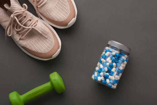 Light-colored sneakers a jar of vitamins in capsules and a green dumbbell on a dark surface