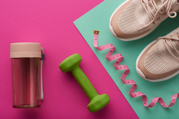 Sports elements such as pink running shoes with a pink ribbon for measurements and a green dumbbell with a sports water bottle are on surfaces of contrasting shades