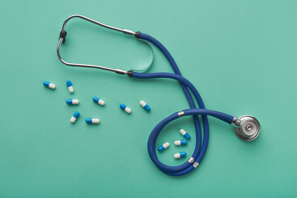 A blue stethoscope with scattered blue-white capsules are on a turquoise surface