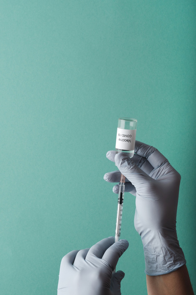 With hands in medical gloves the vaccine is injected into a small syringe with the ampoule turned over against a turquoise background