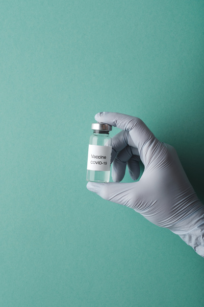 The vaccine in the ampoule is held with a hand in a rubber medical glove on a turquoise background