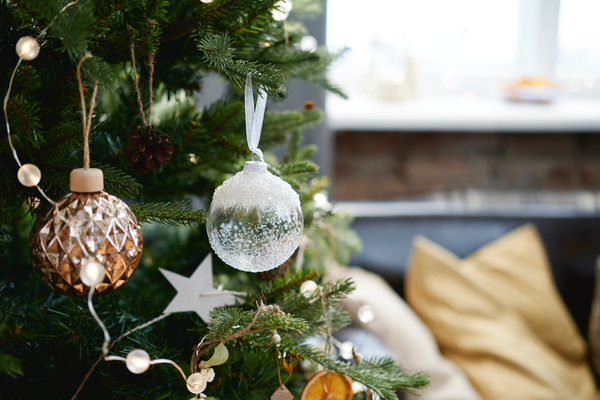 A glass bauble decorated with white sequins imitating frost hangs on the Christmas tree next to a ribbed golden bauble and a garland