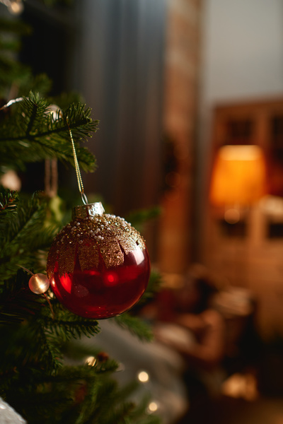 A red glass bauble decorated with golden strokes and beads on a branch of a Christmas tree decorated with a garland in a cozy room with dimmed lights