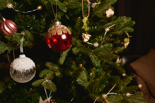 A red bauble decorated with golden strokes and beads hangs next to a glass bauble with white sequins on a decorated Christmas tree with fir cones