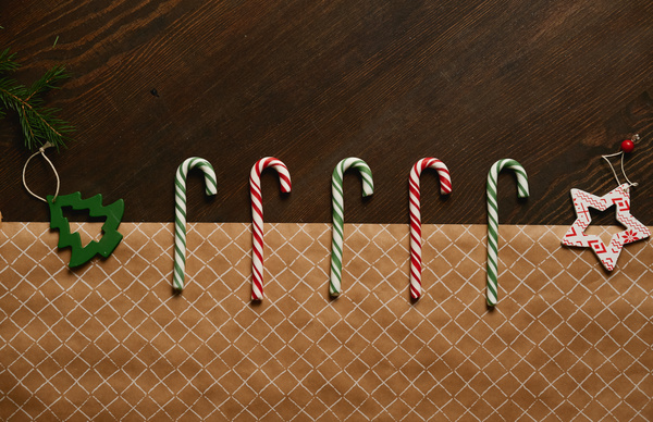 A composition of alternating red and green candy sticks lying with wooden Christmas tree toys on a table made of dark wood covered with craft paper with a pattern