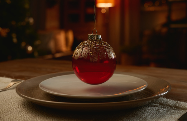 A glass red Christmas bauble with golden strokes and beads suspended above serving plates on a table with a textile napkin and cutlery