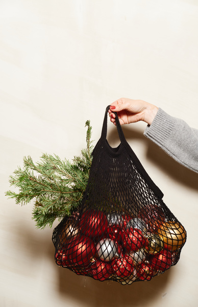 Christmas tree toys of different colors and Christmas tree branches in a black mesh bag held with one hand on a white background