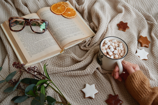 A white mug with a pattern filled with hot chocolate with marshmallows and held by a womans hand on a beige plaid with a book which there are glasses and with glazed gingerbread in the shape of stars