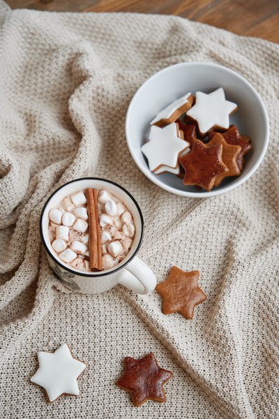 Warming winter drink with marshmallow and cinnamon in a white mug on a blanket with a bowl of gingerbread in a glaze in the form of stars