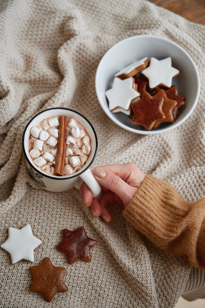 A warming winter drink with marshmallows and cinnamon in a white mug held by a womans hand on a blanket and a bowl of gingerbread in a glaze in the form of stars