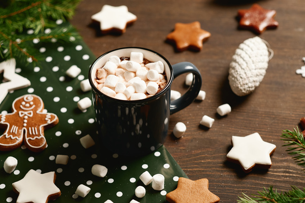 Cocoa with marshmallows in a dark gray mug on a table with green polka dot wrapping paper scattered gingerbread of different shapes Christmas tree branches and toys