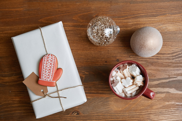 Cocoa with marshmallow in a burgundy mug next to a Christmas gift in a white package tied with string with glazed gingerbread in the shape of a glove and Christmas tree shining baubles
