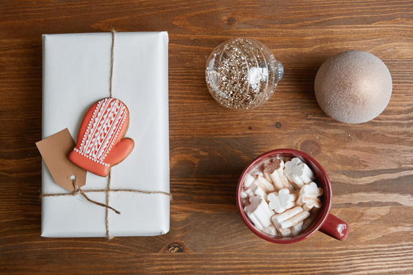 Hot winter drink with marshmallows in dark red mug with a Christmas gift in a white package tied with string with gingerbread in the shape of a glove and Christmas tree shining baubles on a dark wood table