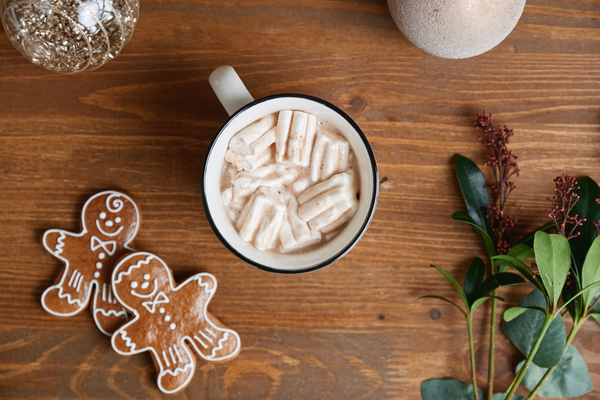 A mug of hot winter drink with marshmallows on a wooden table with gingerbread men and shiny Christmas baubles