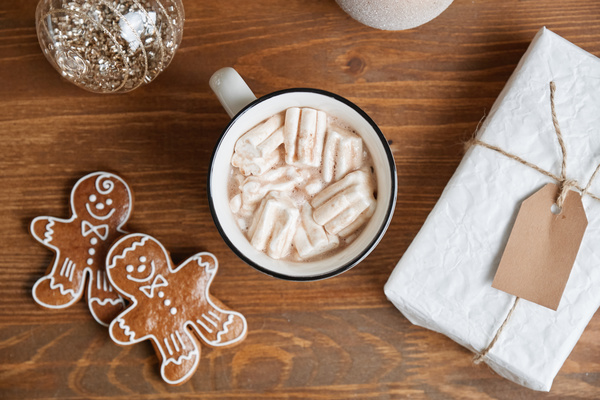 A mug of cocoa with marshmallows with gingerbread men a Christmas gift in white wrapping paper and shiny Christmas baubles on a wooden table