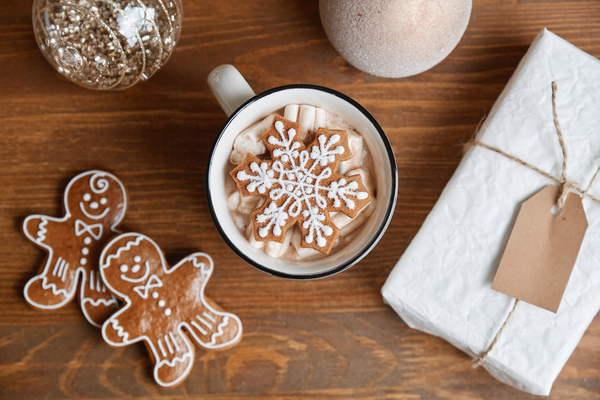 Cocoa with gingerbread snowflake on a wooden table with gingerbread men and a Christmas gift in white wrapping paper near with shiny Christmas baubles