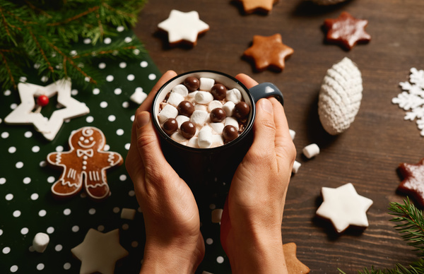 A Christmas drink with marshmallows and chocolate balls in a mug held with two hands on a table with gingerbread tree branches and toys