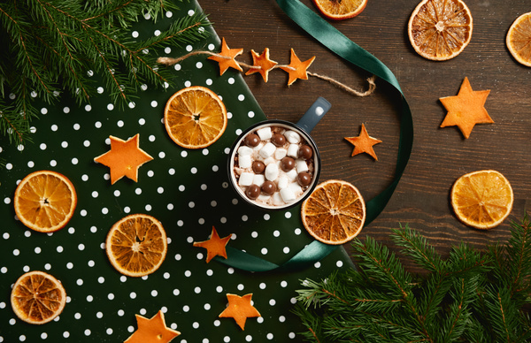 A mug of cocoa with chocolate balls on the table with a scattering of dried orange slices stars from the peel and with fir branches and a green ribbon