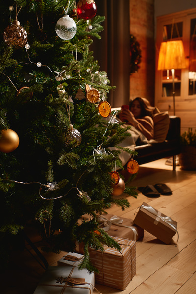 A decorated Christmas tree with gifts on the background of which a woman resting on the sofa on Christmas evening in a room with a dim light