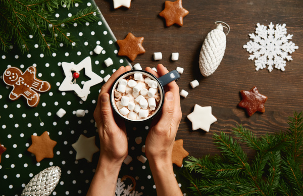 A mug filled with cocoa with marshmallows in his hands against the background of Christmas tree branches toys and gingerbread of different shapes