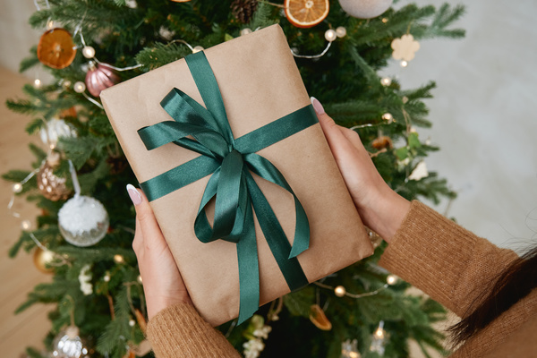 A Christmas present wrapped in craft paper and decorated with a dark green ribbon in female hands on the background of a decorated Christmas tree