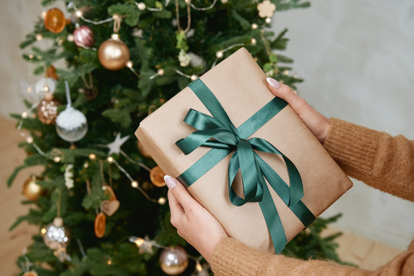 A Christmas gift wrapped in craft paper and tied with a dark green ribbon in womens hands with a decorated Christmas tree on the background