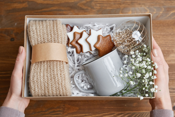 A Christmas gift box with a mug a Christmas bauble and a knitted scarf held in the hands