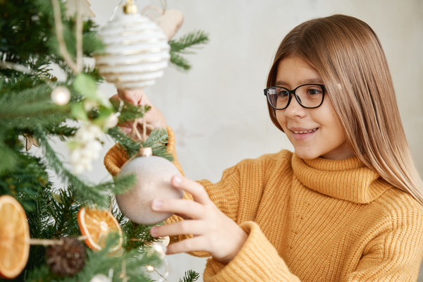 A girl with brown hair in black glasses and a yellow sweater hangs a light colored Christmas bauble on the Christmas tree