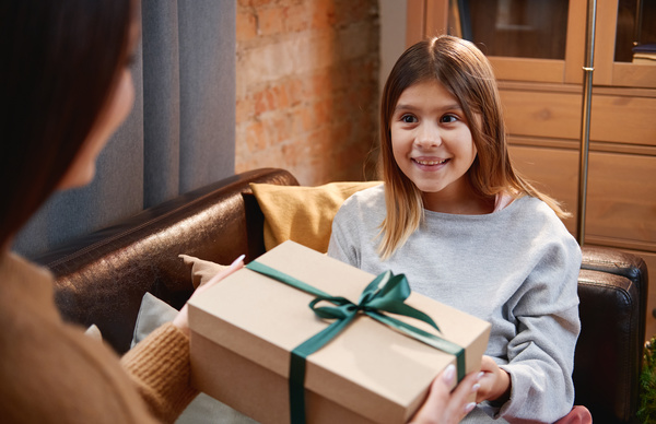 A daughter with brown hair with joy on her face receives a Christmas gift from her mother in a box tied with a green satin ribbon sitting on the sofa